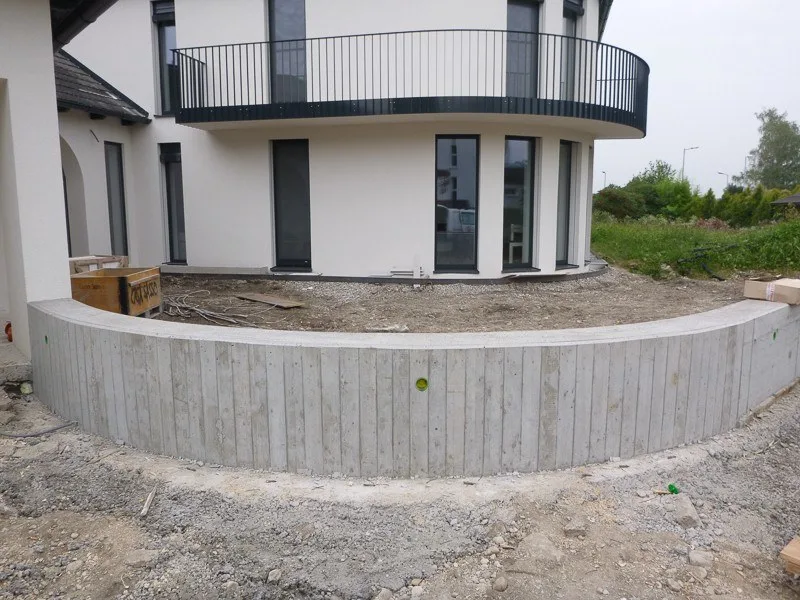 Runde Sichtbetonmauer vor einem neuen weißen Wohnhaus mit Balkon