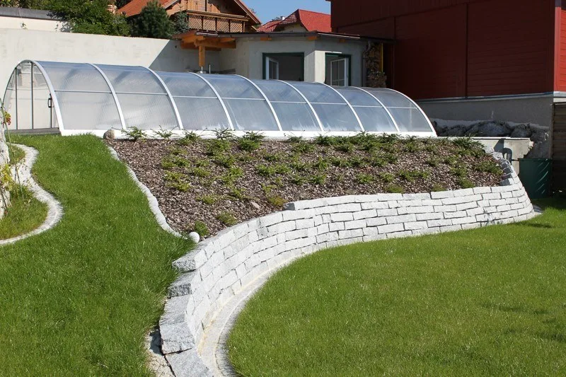 Geschwungene Natursteinmauer im Garten vor einer Poolüberdachung